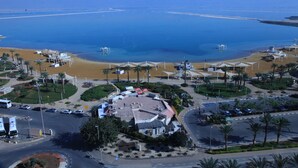 An aerial view of a coastal resort with a beach, palm trees, and a central building with a red roof.