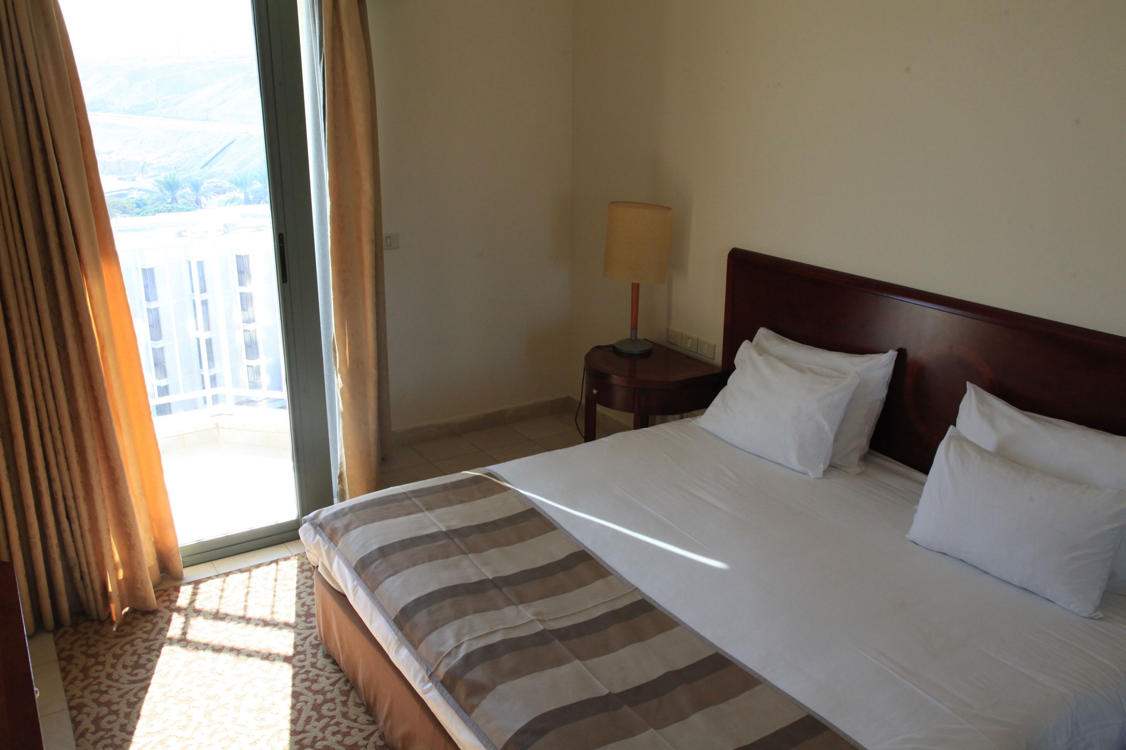 Habitación de hotel con una cama grande, una mesita de noche con lámpara y vistas al exterior a través de una puerta corrediza.