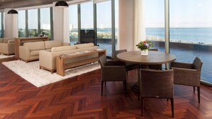 A spacious living room with a large window overlooking the ocean, a round dining table with chairs, and a sofa set.