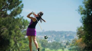 A golfer in mid-swing on a golf course with a clear sky and trees in the background.
