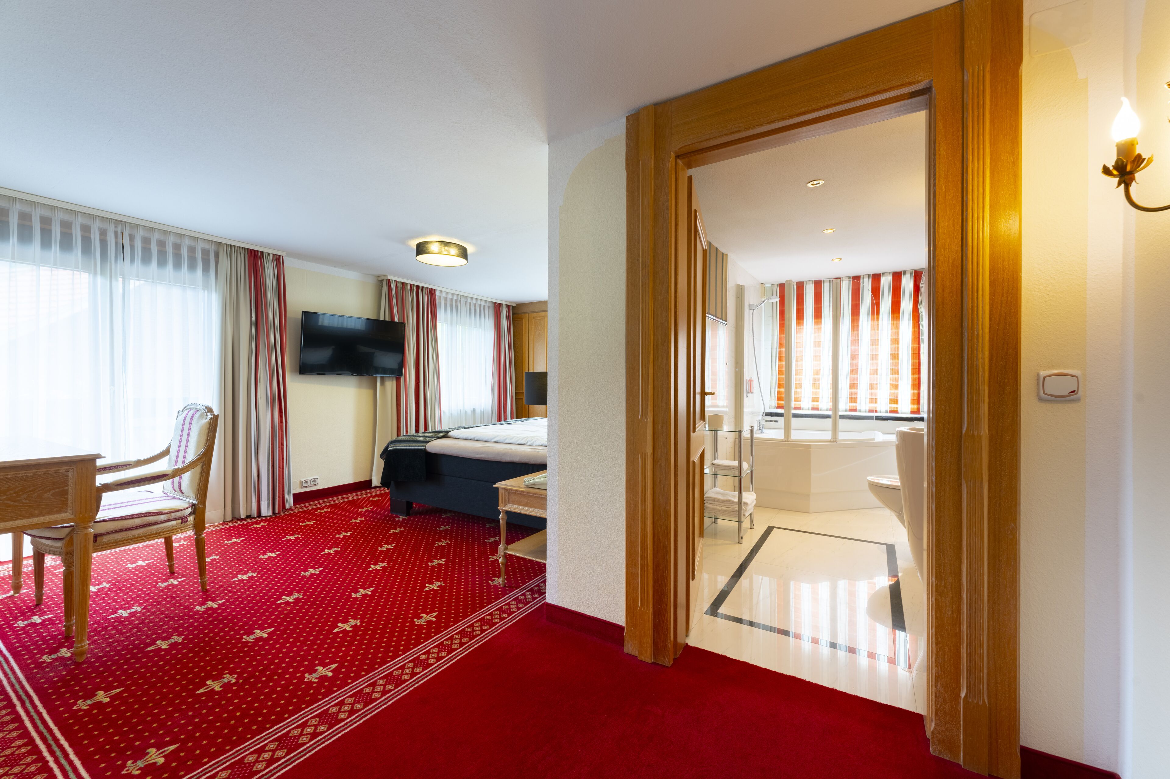 Habitación de hotel con cama, escritorio, silla, televisor y baño visible a través de una puerta abierta.