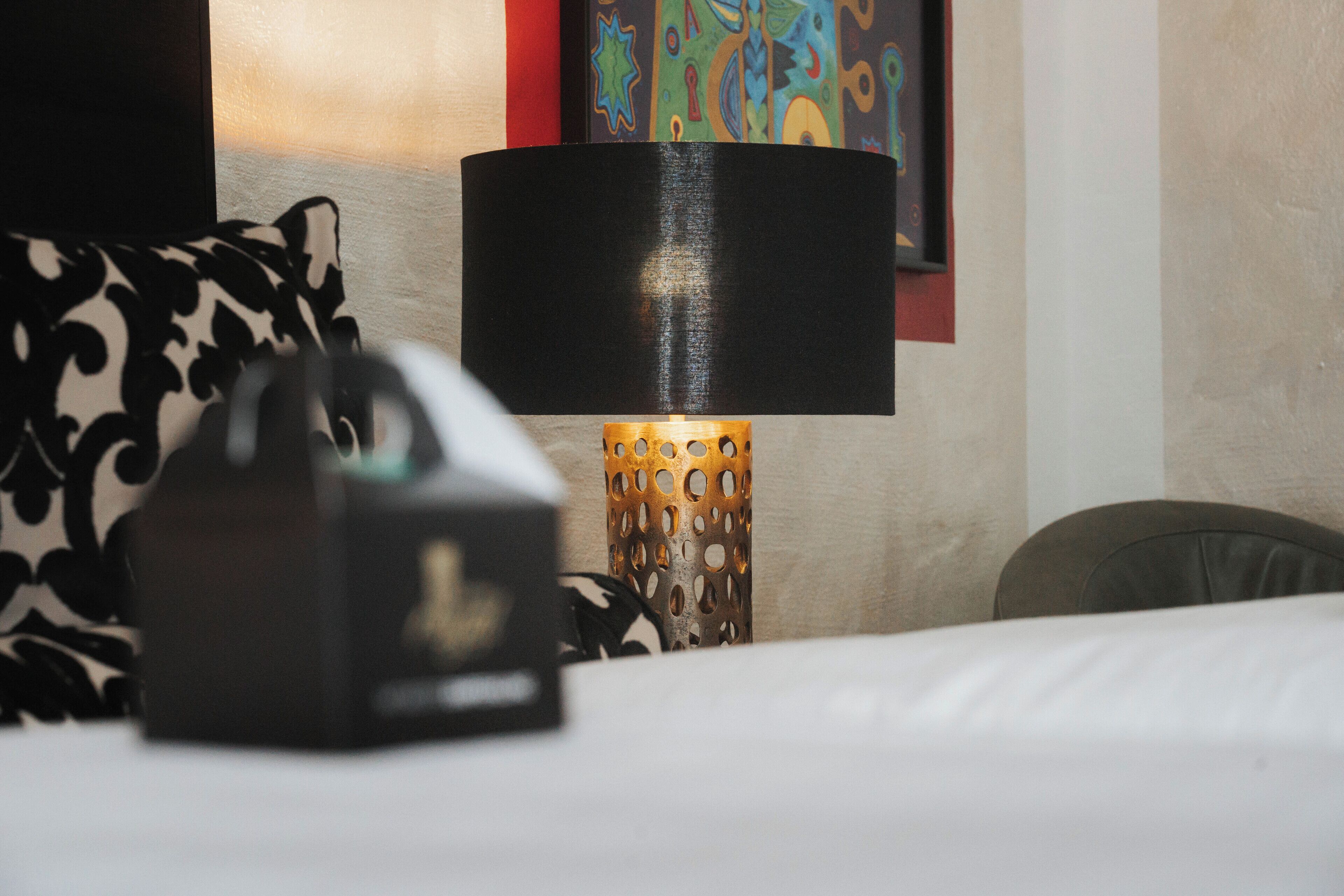 Une chambre d’hôtel avec un lit, un oreiller à motifs noir et blanc, une lampe avec un abat-jour ajouré et un tableau coloré accroché au mur.