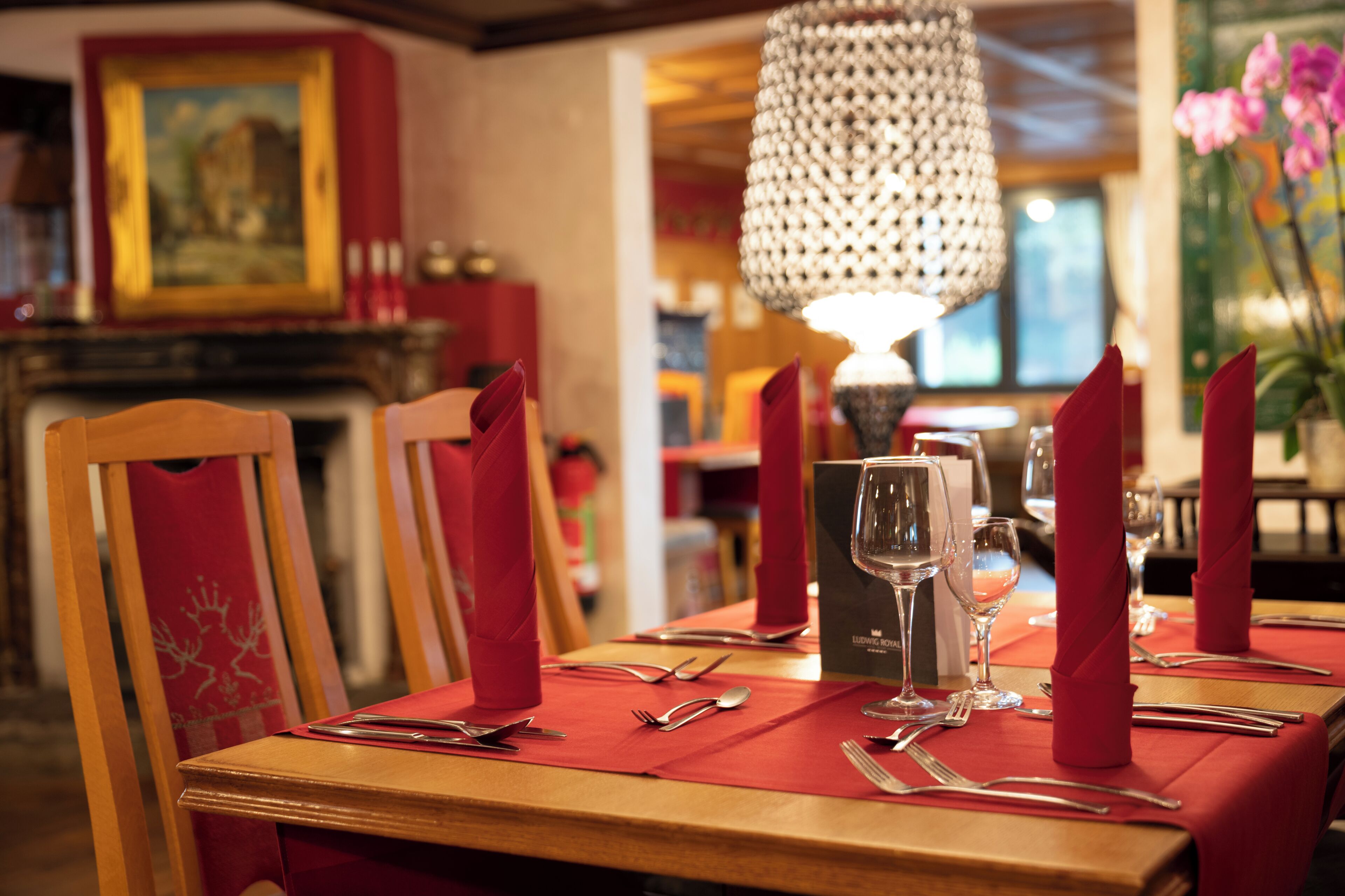 Una mesa de comedor dispuesta con servilletas rojas, cubiertos y copas de vino, con una araña y un cuadro al fondo.