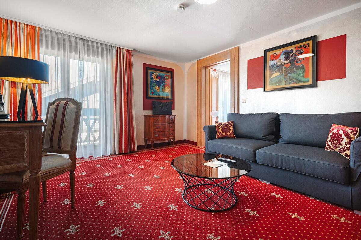 A hotel room with a red patterned carpet, a blue sofa, a wooden chair, a glass coffee table, and a painting on the wall.