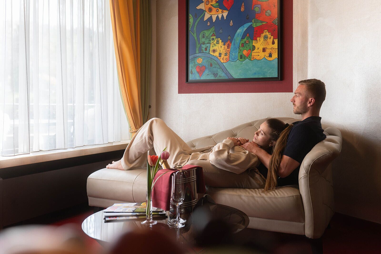 Ein Paar entspannt auf einem beigen Sofa in einem gut beleuchteten Raum mit einem großen Fenster, einem Bild an der Wand und einem kleinen Tisch mit einer Vase und einem Glas.