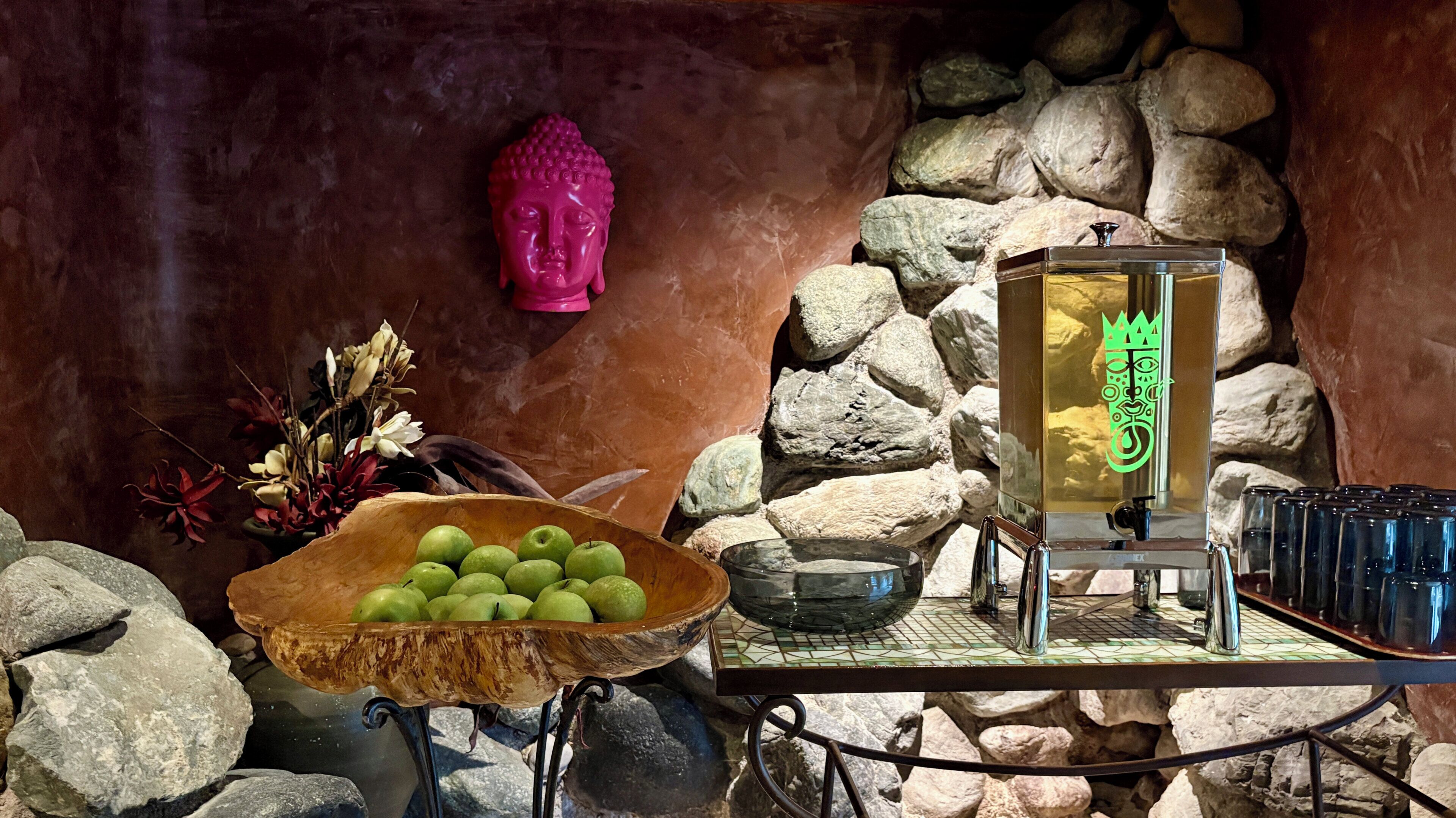 A stone wall with a Buddha statue, a bowl of green apples, and a water dispenser.
