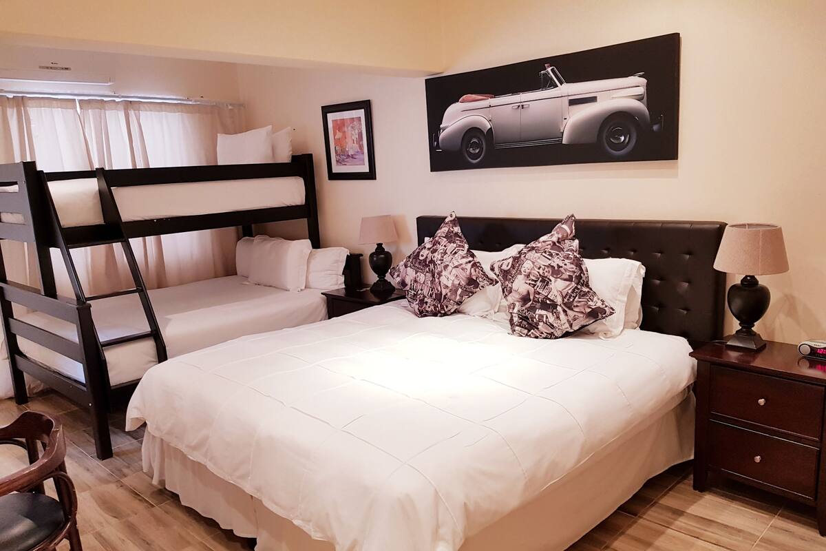 A hotel room with a bunk bed, a large bed with patterned pillows, a nightstand with a lamp, and a framed picture on the wall.