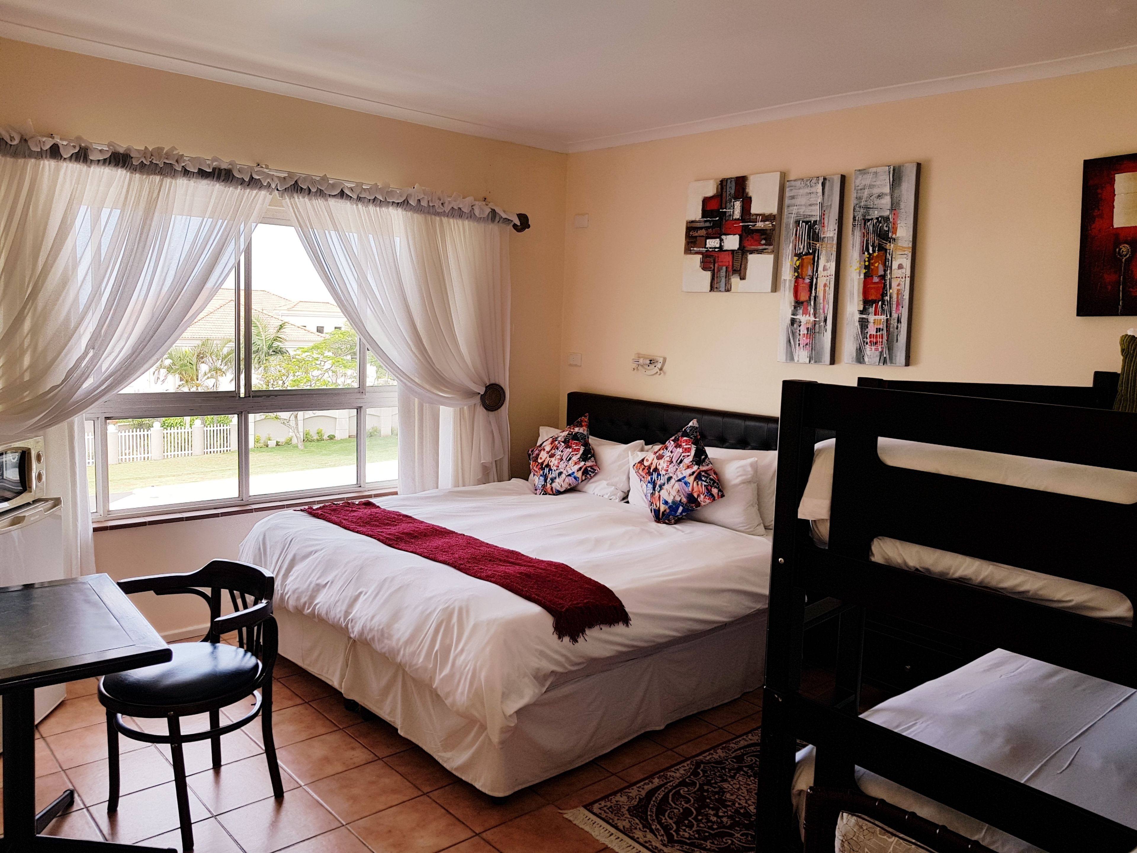 A bedroom with a bed, a desk, and a chair. There is a window with curtains, a wall with artwork, and a view of the outside.