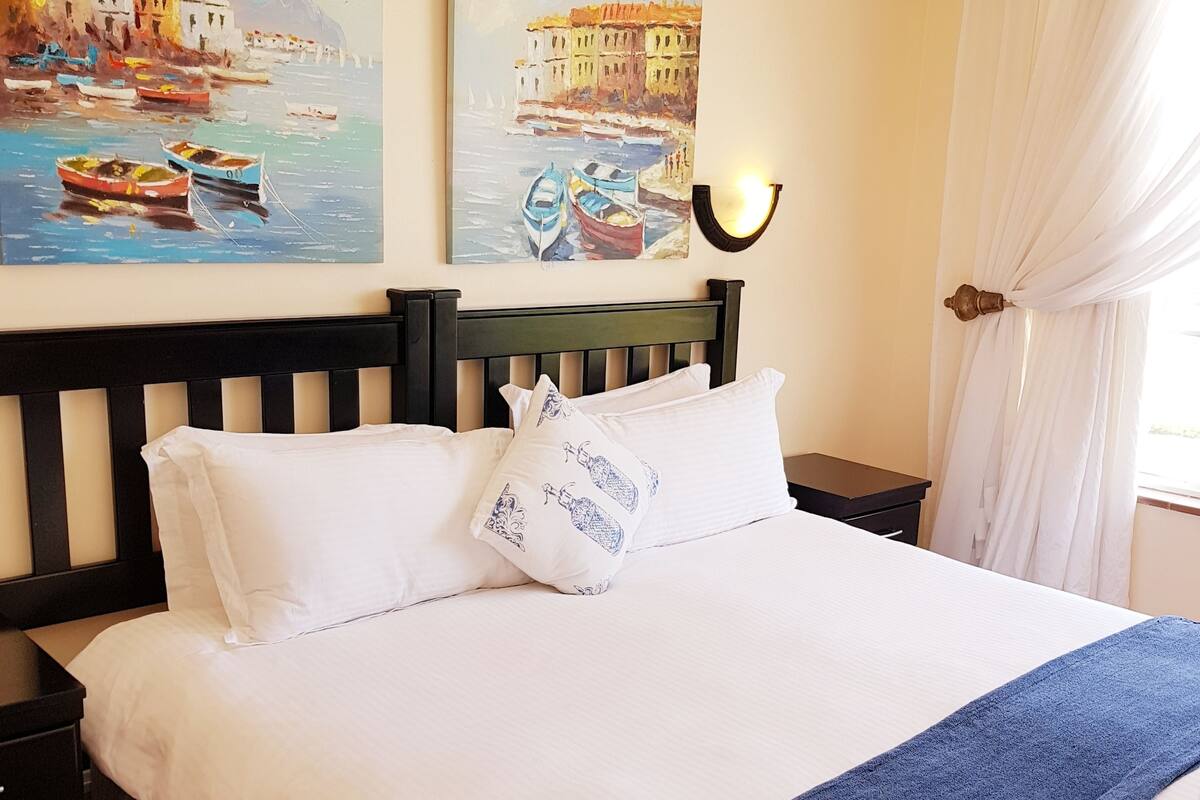 A neatly made bed with white linens and a blue throw blanket, a black headboard, and two paintings depicting coastal scenes.