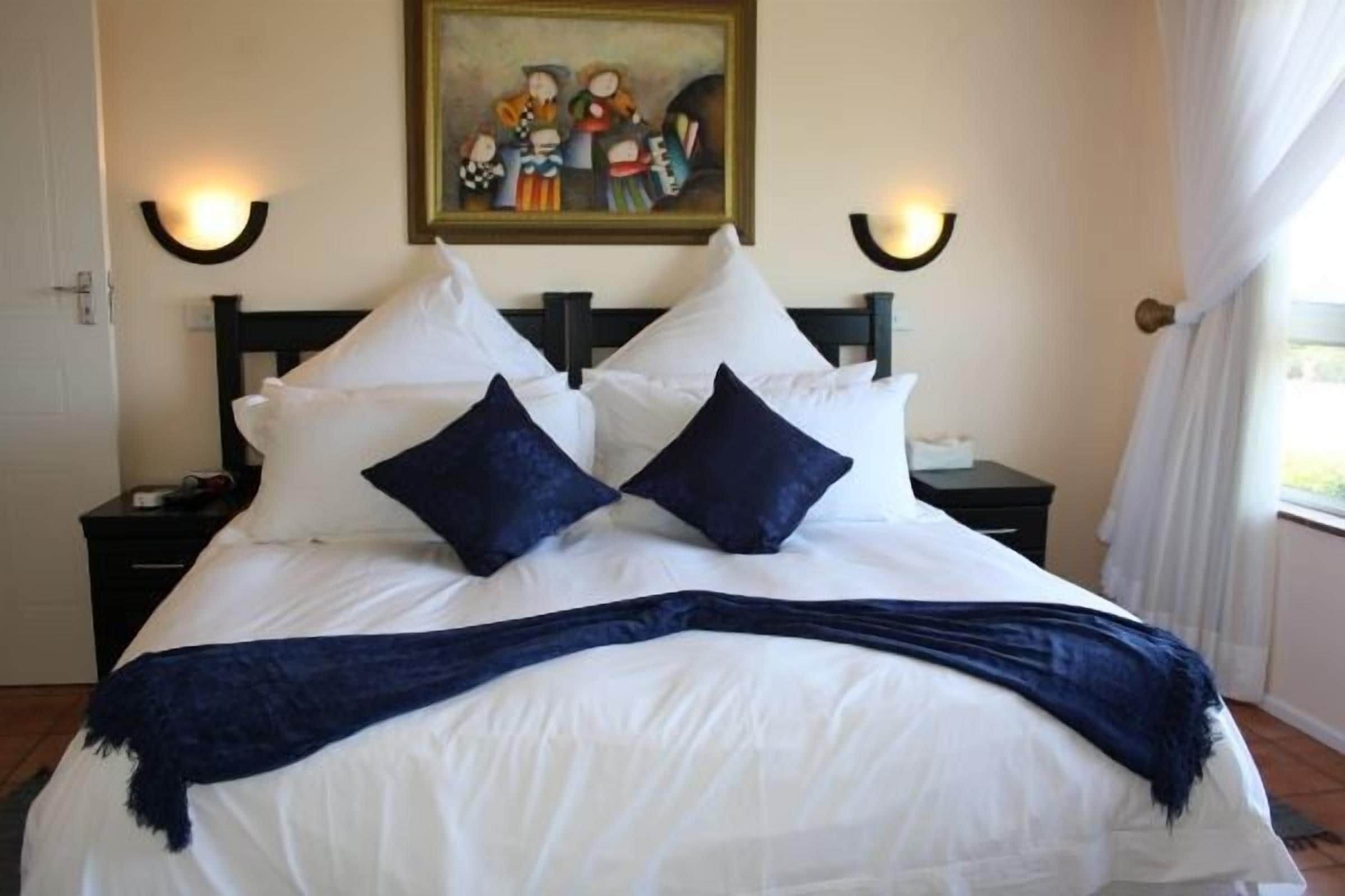 A neatly made bed with white linens and two dark blue pillows, a painting above the bed, and wall-mounted lights on either side.