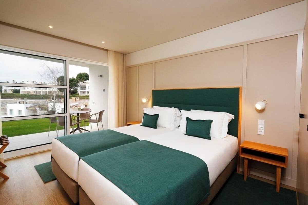 Ein Hotelzimmer mit einem großen Bett, einem kleinen Tisch, einem Sessel und Blick auf ein Gebäude sowie ein Gewässer.