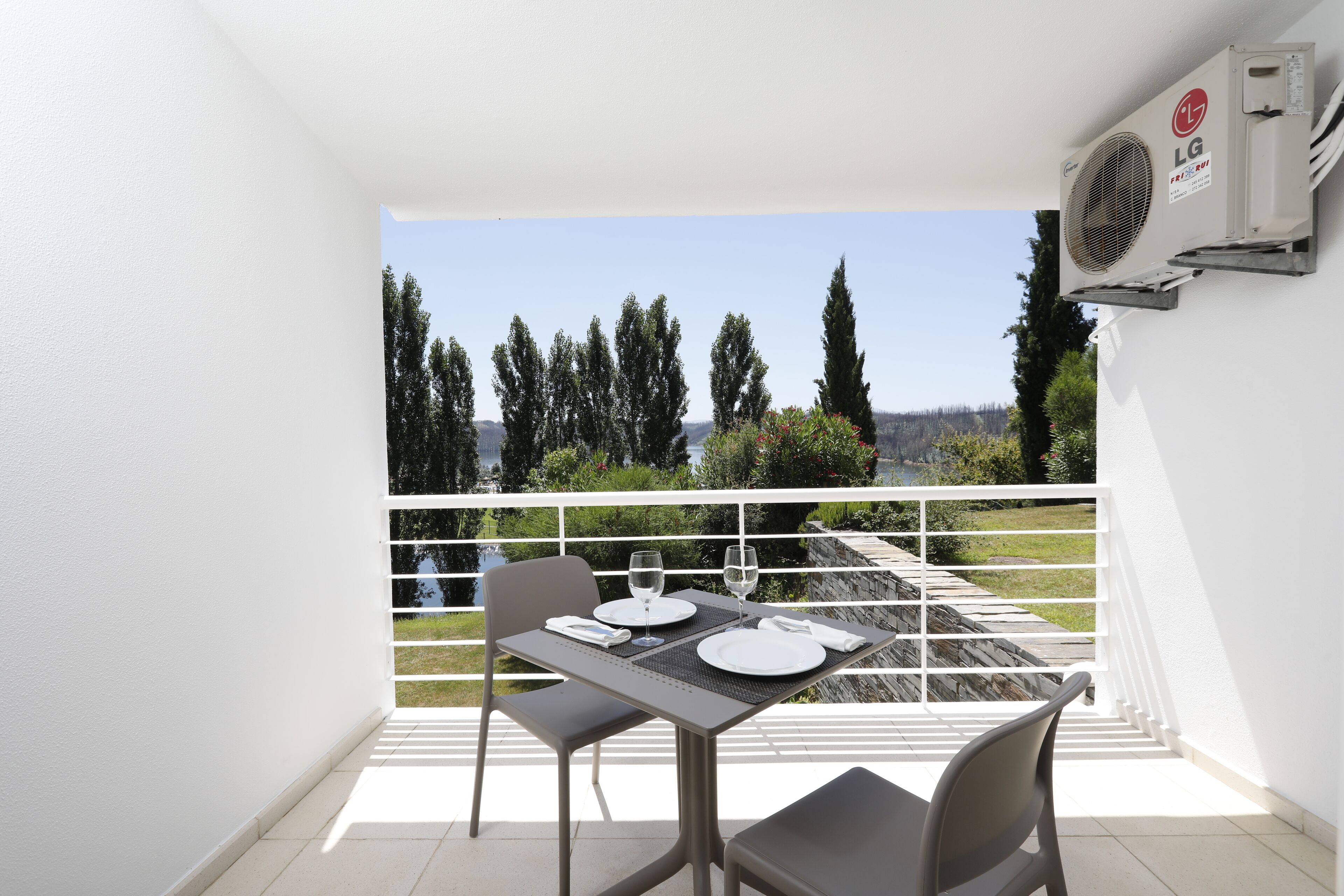 A balcony with a table set for two, overlooking a scenic landscape with trees and a body of water.