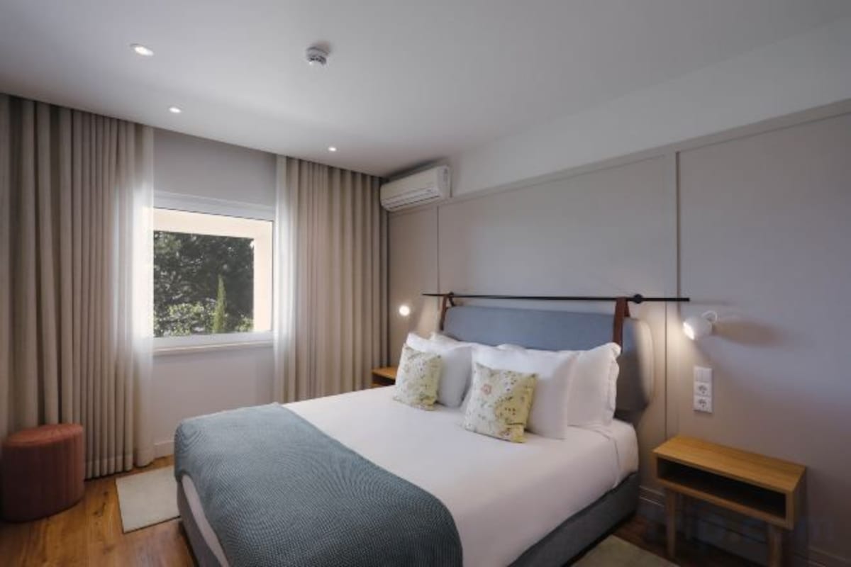 A modern hotel room with a large bed, a bedside table, a window with curtains, and an air conditioning unit.