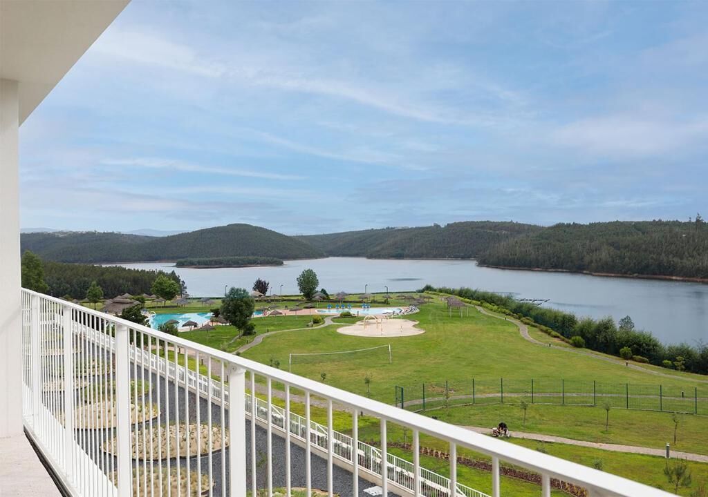 Un balcón con vistas a un lago, zonas verdes y un parque infantil.