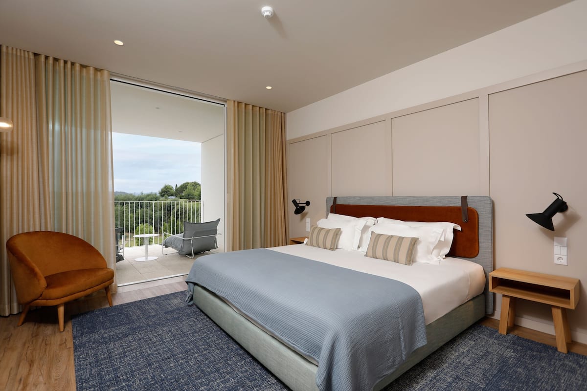 Ein modernes Hotelzimmer mit einem großen Bett, einem Sessel, einem kleinen Tisch und einem Balkon mit Blick.