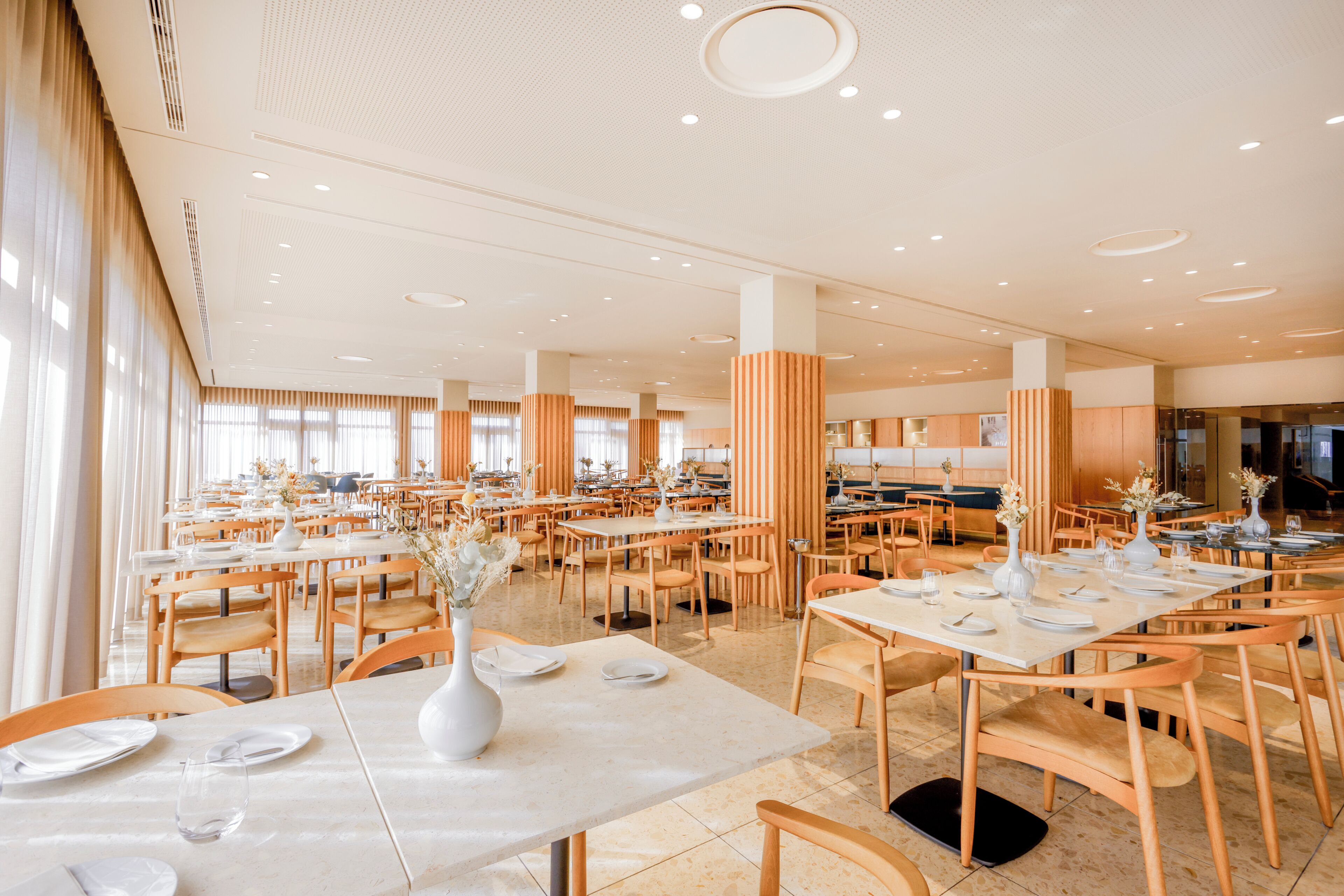A spacious dining area with multiple tables and chairs, large windows, and a high ceiling with recessed lighting.
