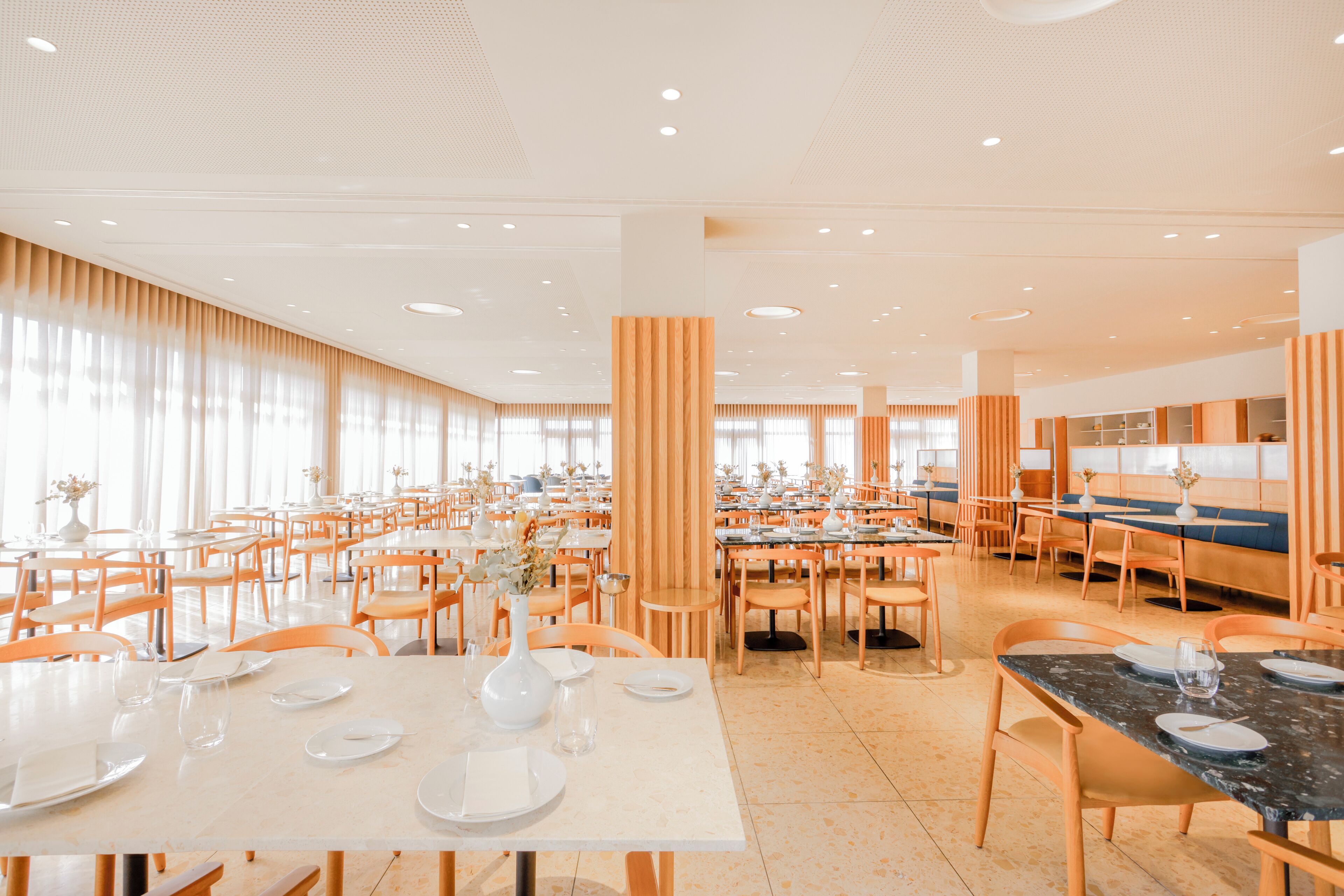 Un comedor espacioso con varias mesas y sillas, ventanas grandes con cortinas y un diseño moderno en el techo.
