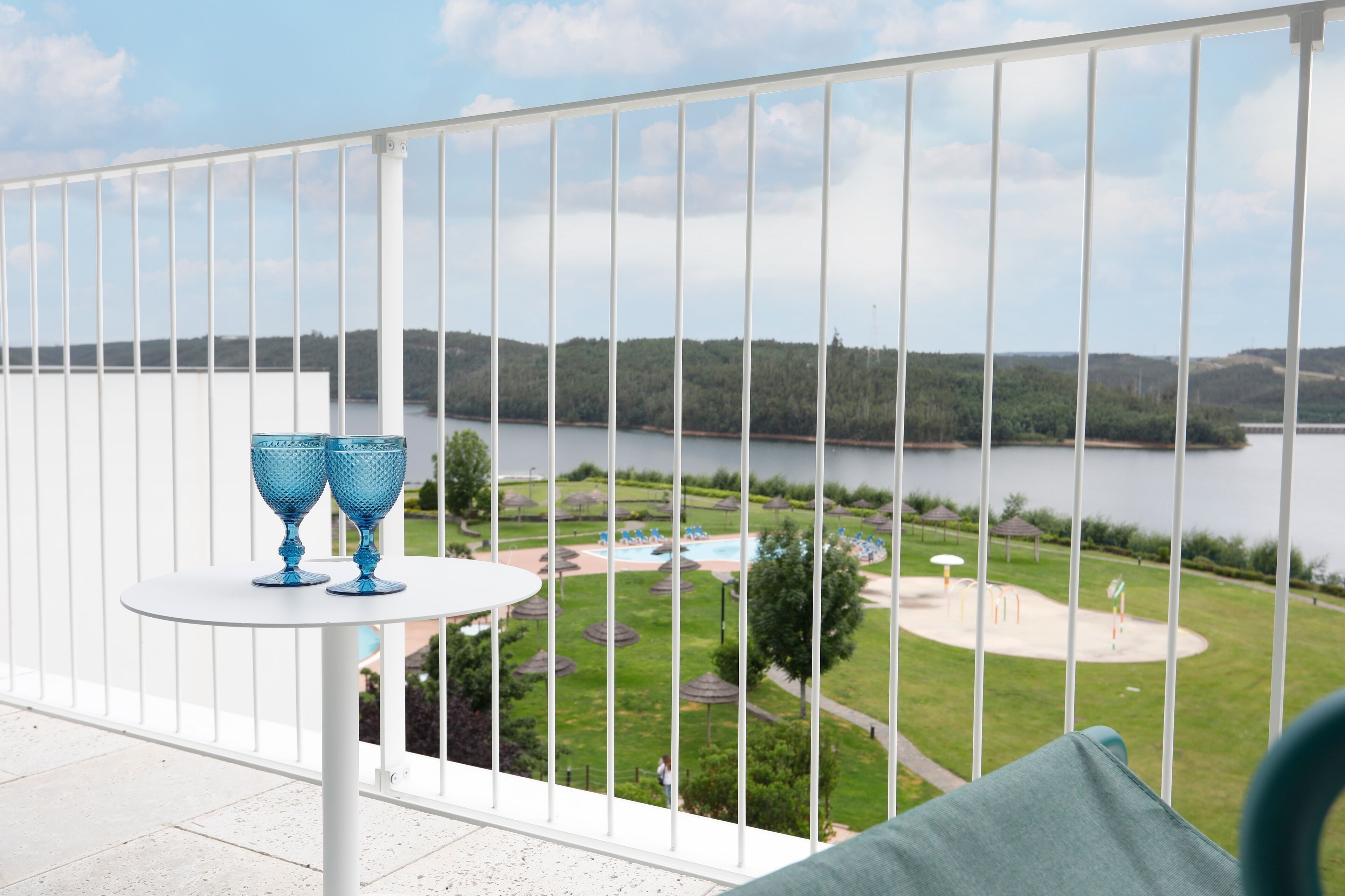 En balkong med ett vitt räcke, två blå glasvaser på ett litet runt bord och utsikt över en sjö och ett parkområde med träd och en lekplats.