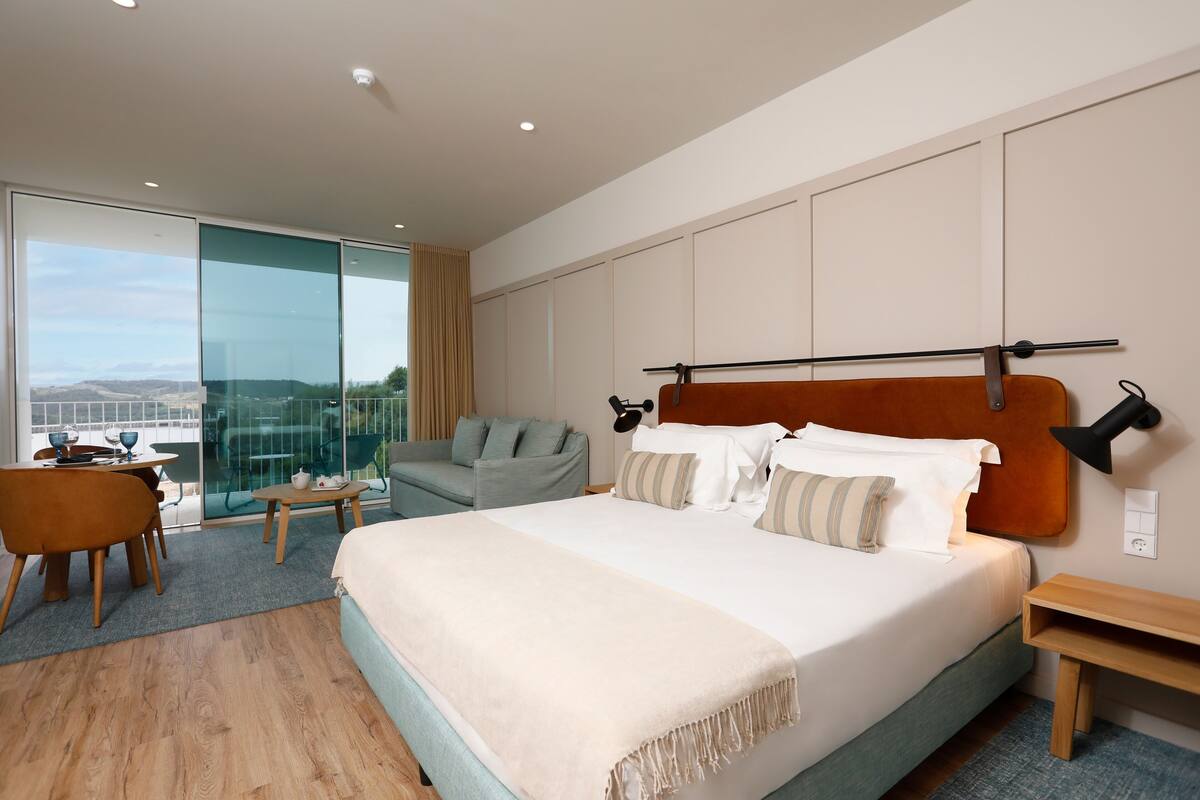 Ein modernes Hotelzimmer mit einem großen Bett, einer Sitzecke mit Tisch und Stühlen, einem Sofa und Blick durch eine Glastür.