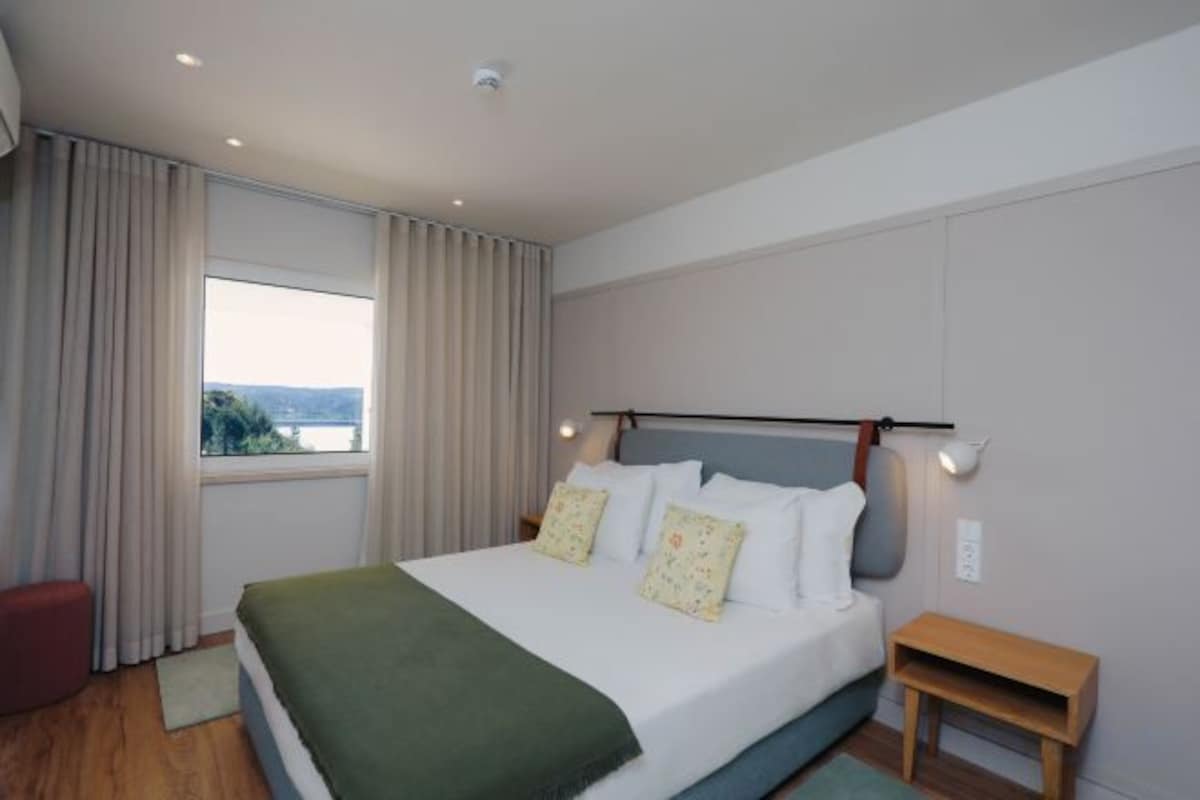 A modern hotel room with a large bed, a view of the outdoors, and a small wooden side table.