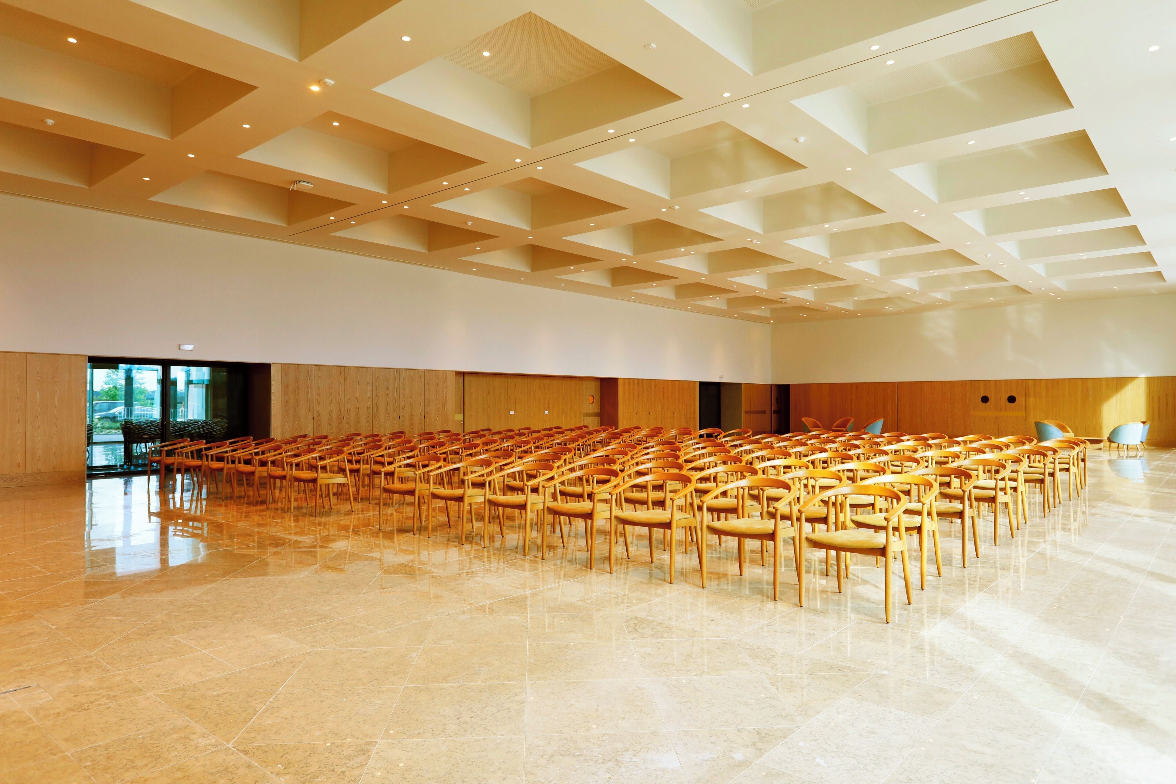 A spacious hall with rows of wooden chairs, a high ceiling with recessed lighting, and a polished floor.