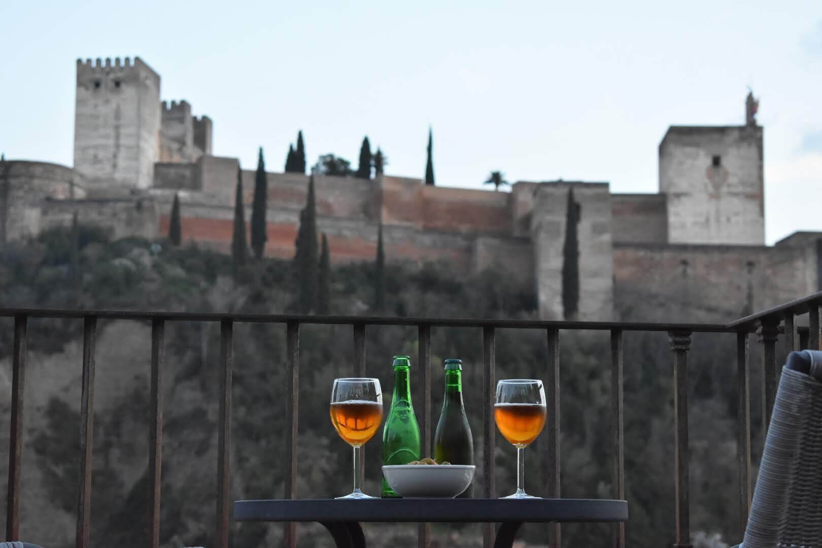 A balcony with a table set for two, featuring two glasses of amber liquid and two green bottles, overlooking a historic stone wall with towers.