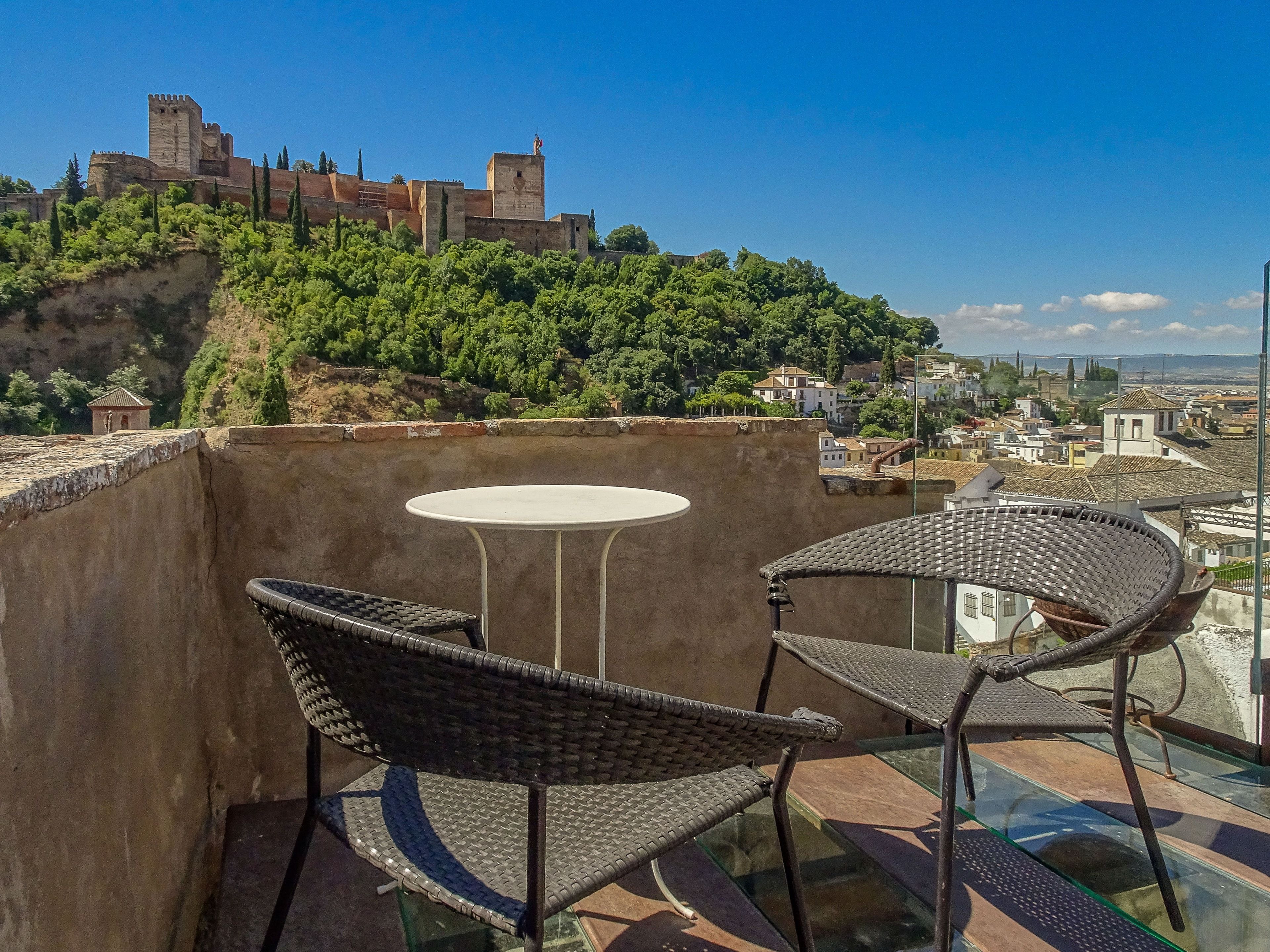 A balcony with wicker chairs and a small round table, overlooking a historic castle and a town.