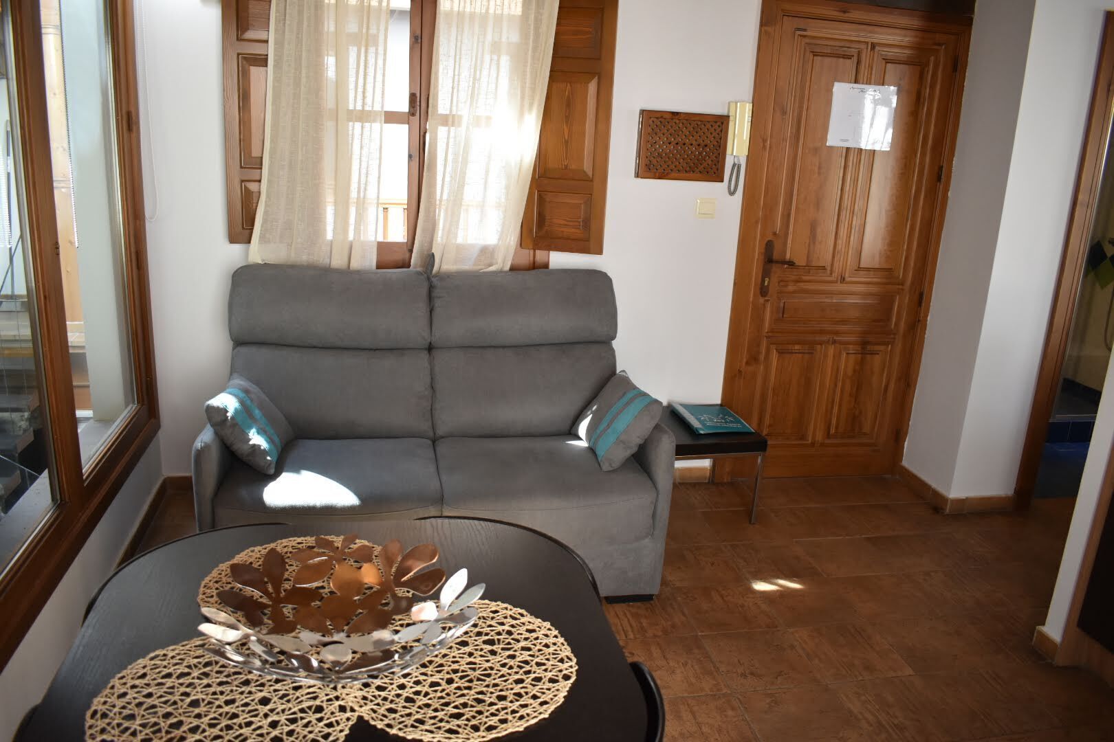 Ein Wohnzimmer mit grauem Sofa, einem kleinen Tisch mit dekorativer Schale und einer Holztür im Hintergrund.