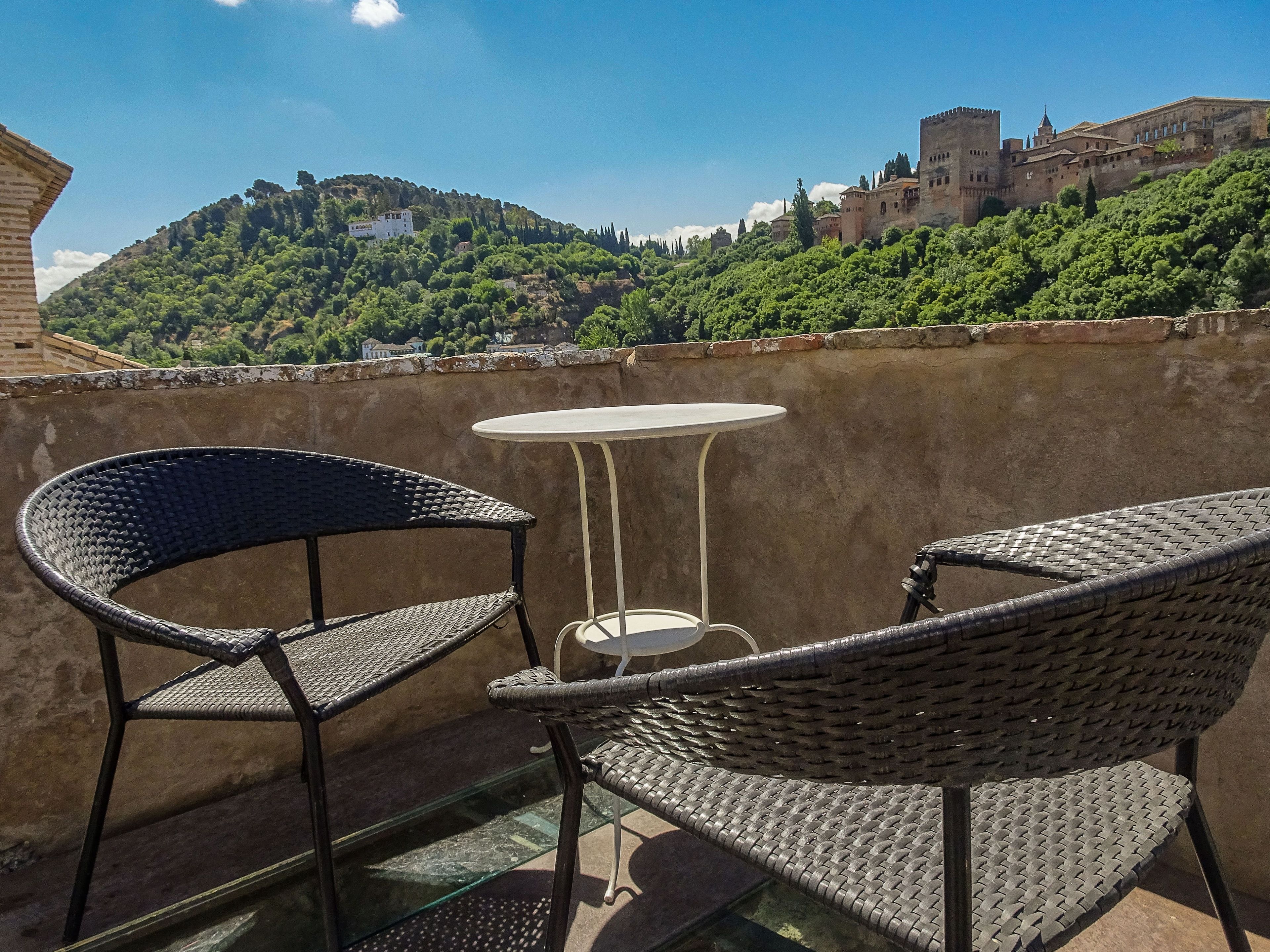 Un balcón con sillas de mimbre y una pequeña mesa redonda, con vistas a un castillo histórico y colinas verdes exuberantes.