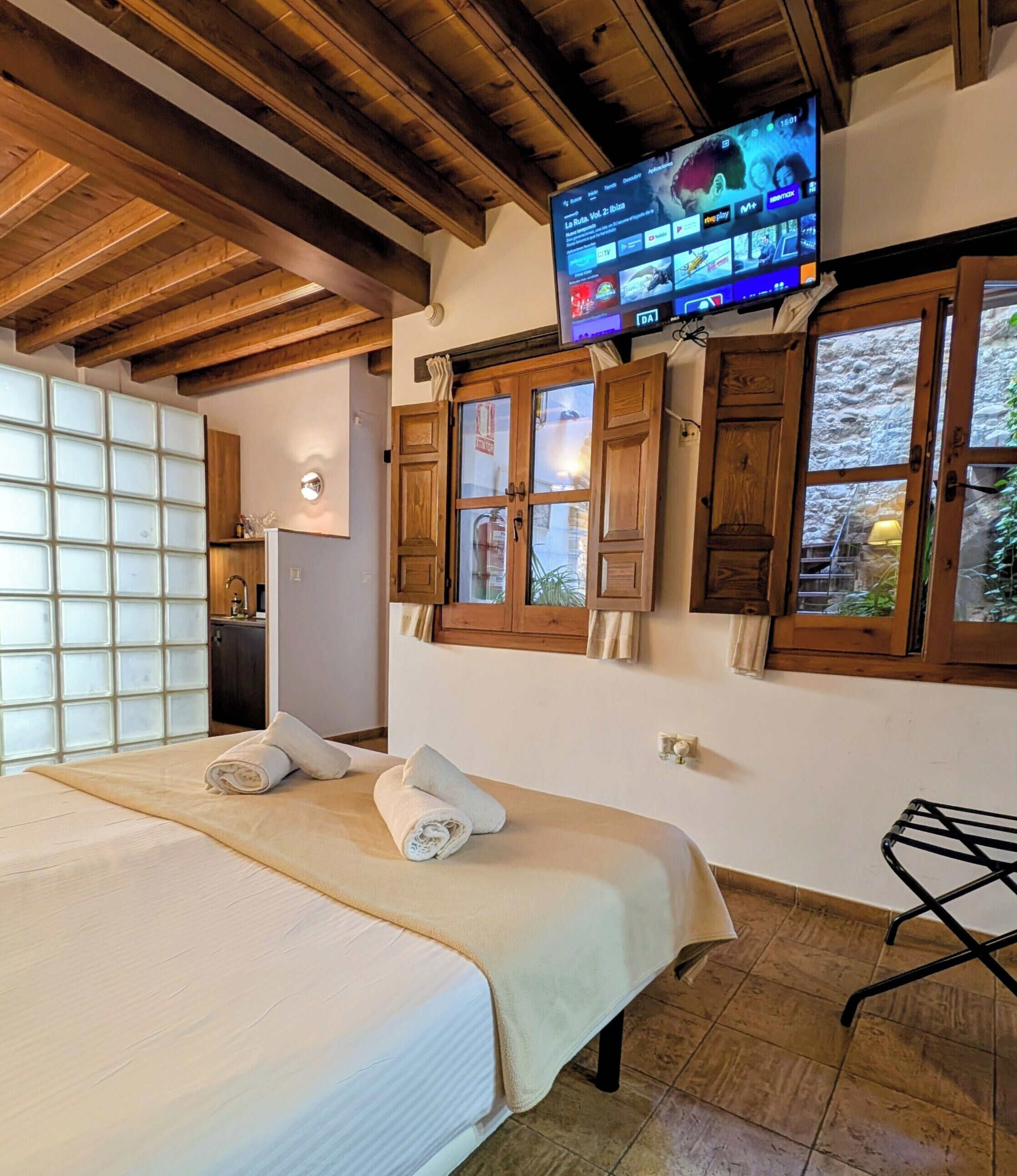 A hotel room with a bed, a television mounted above a window, and wooden beams on the ceiling.