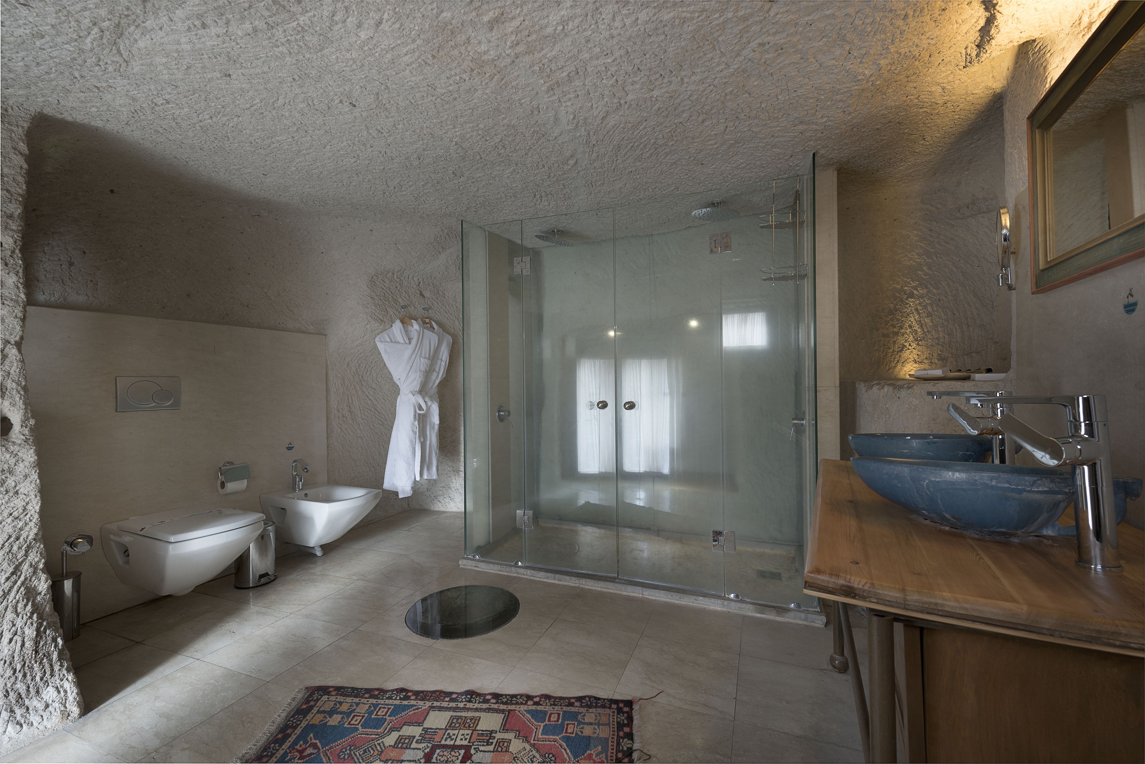 A bathroom with a glass-enclosed shower, a white toilet, a bidet, and a wooden vanity with a blue basin.