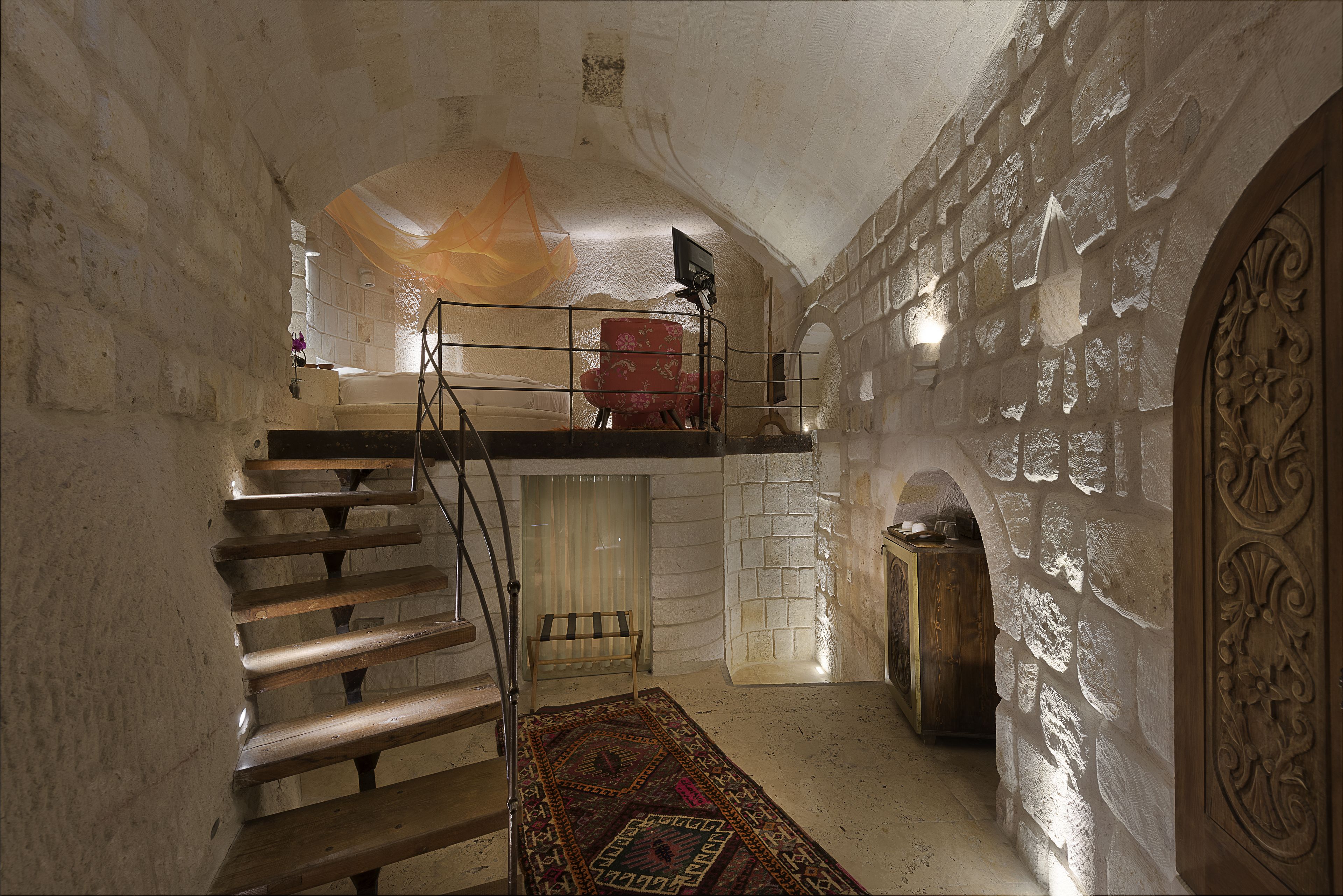 Ein Zimmer mit Steinwänden, einer Holztreppe, einem roten Sessel und einem gemusterten Teppich.