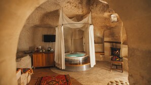 A cozy, circular room with a stone ceiling, a round bathtub, a wooden cabinet, and a small table with a fire. - Anatolian Houses Cave Hotel & SPA (Nevsehir)
