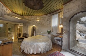 A bedroom with a stone wall, a large bed, a wooden desk, and a view of a rocky landscape through an arched doorway. - Anatolian Houses Cave Hotel & SPA (Nevsehir)