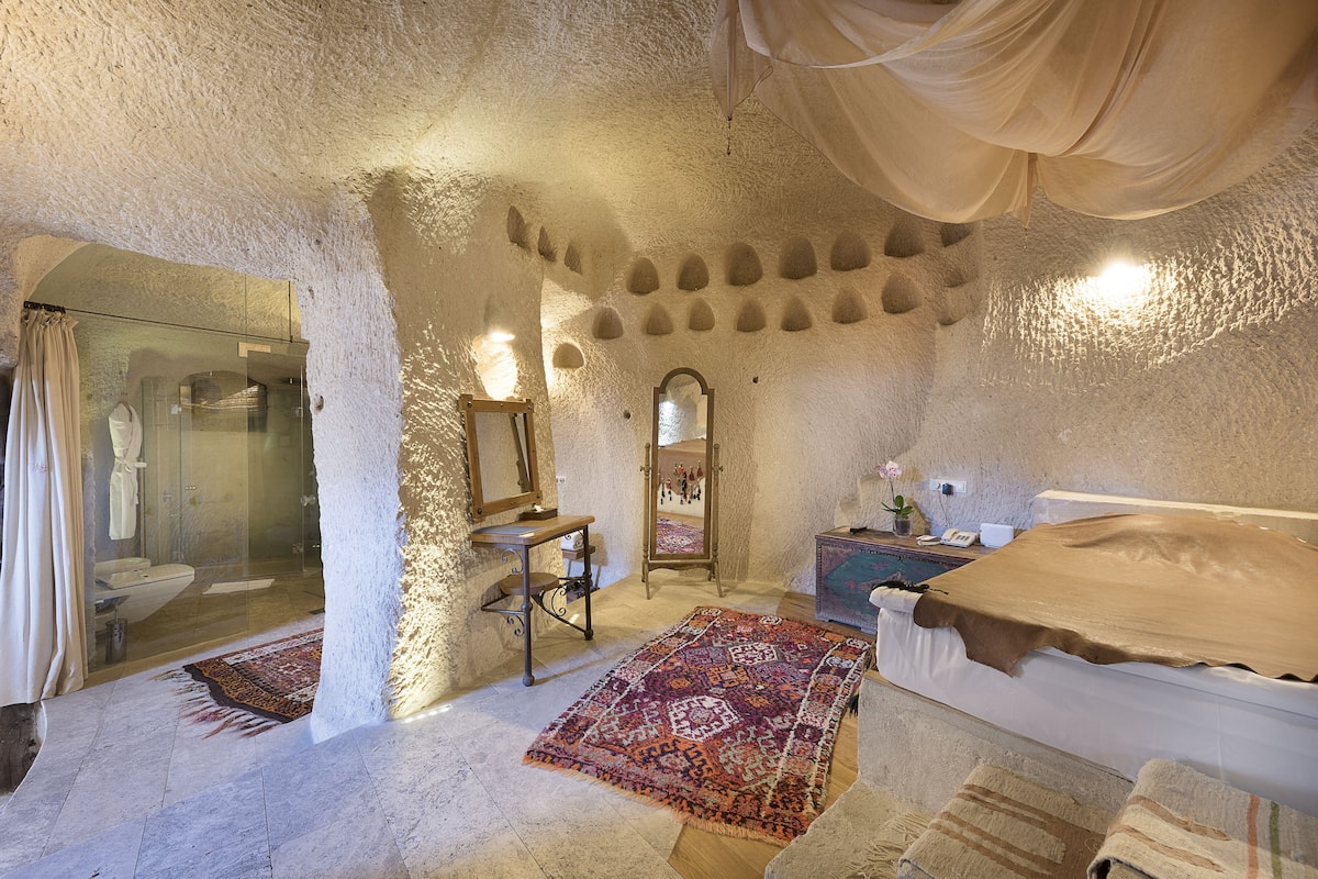 A room with a stone wall, a bed with a brown cover, a mirror, a wooden cabinet, and a patterned rug.