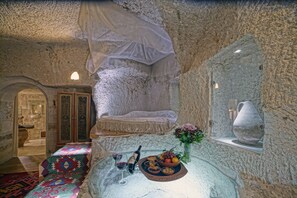 A stone-walled room with a bed, a glass table with a bottle, glasses, and a fruit bowl.