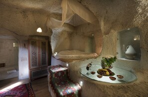 A room with a stone wall, a bed with a canopy, a glass table with fruit and a vase, and a patterned bench. - Anatolian Houses Cave Hotel & SPA (Nevsehir)