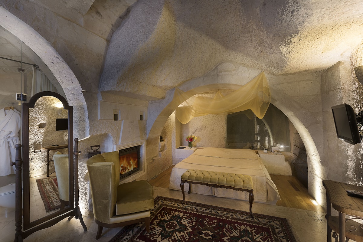 A bedroom with a bed, a chair, a mirror, a fireplace, and a television.