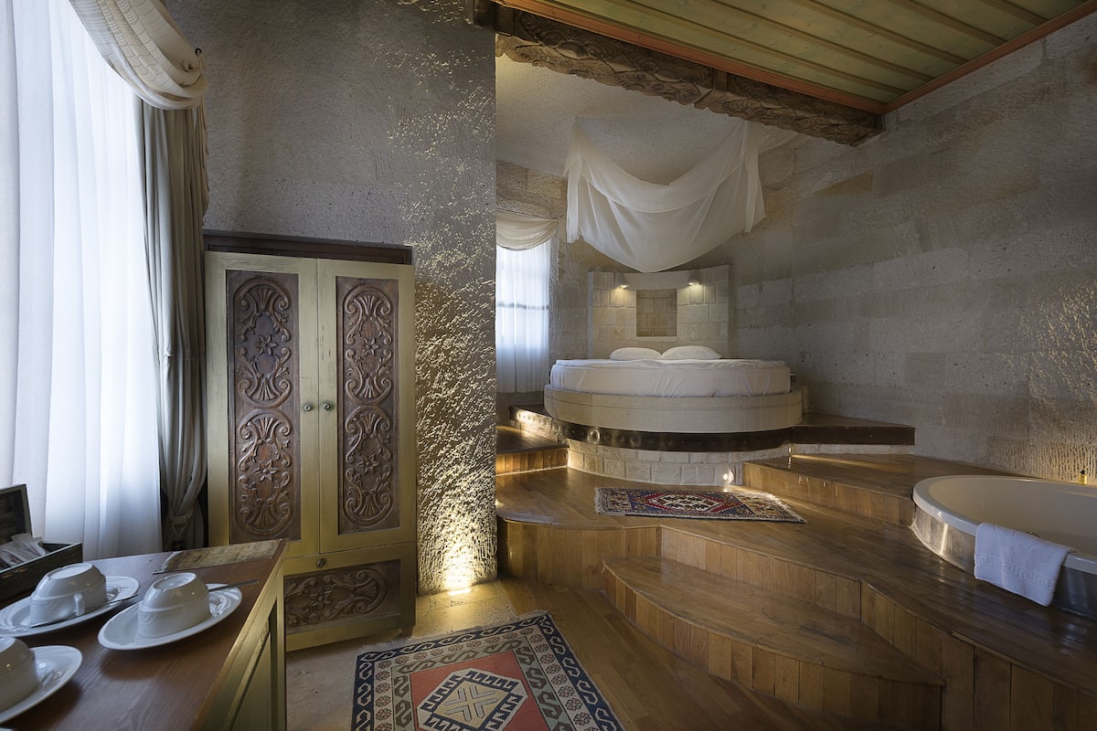 A room with a stone wall, a wooden floor, a bed with a white canopy, a bathtub, and a table with plates.