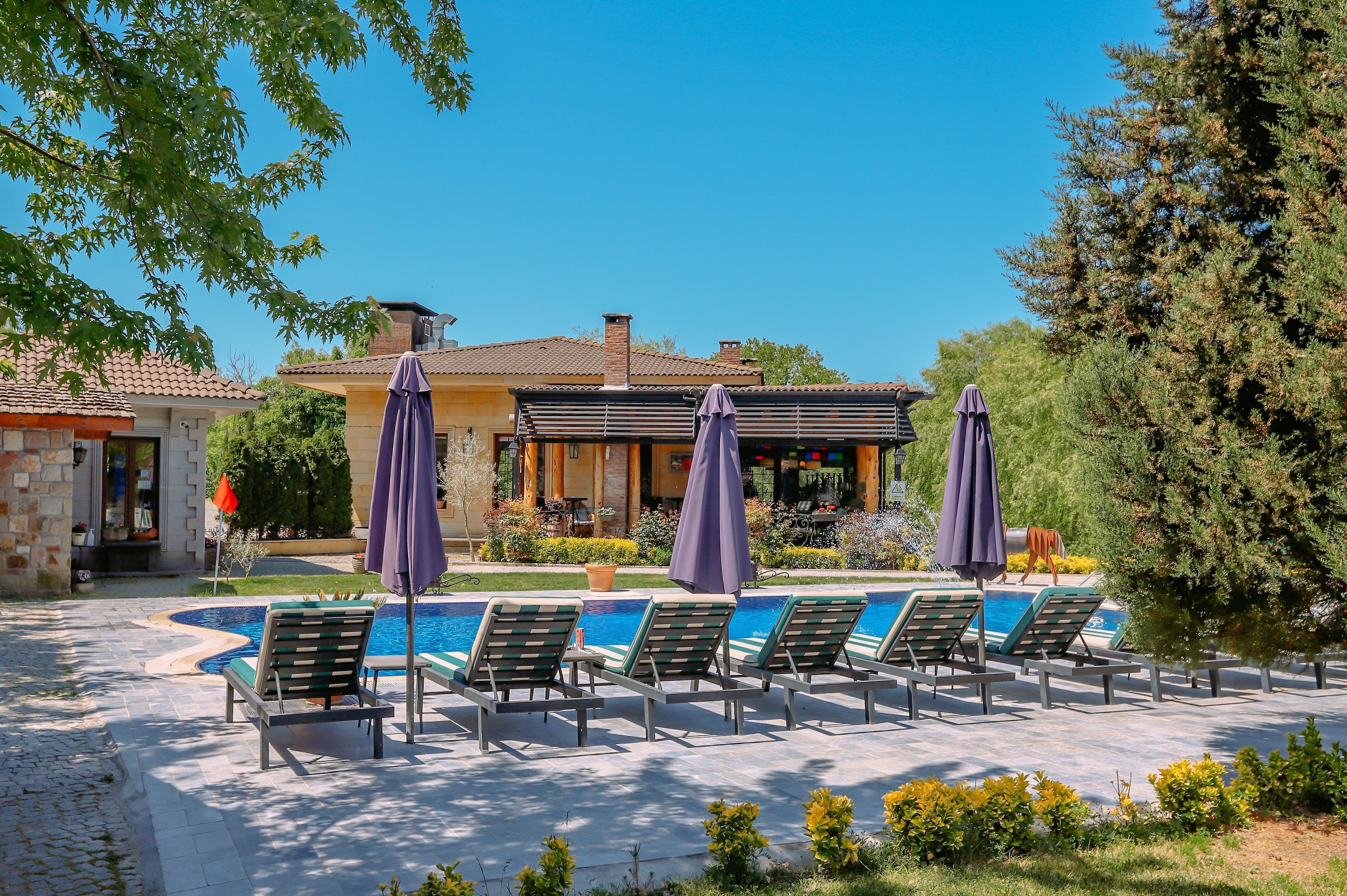 Un área de piscina con sillones de descanso y sombrillas, un edificio con tejado de baldosas y un jardín bien cuidado.