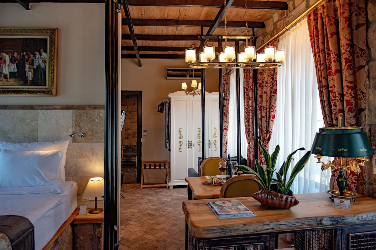 Quarto de hotel com uma cama, uma mesa com abajur, uma cadeira, um vaso de planta e um quadro na parede.