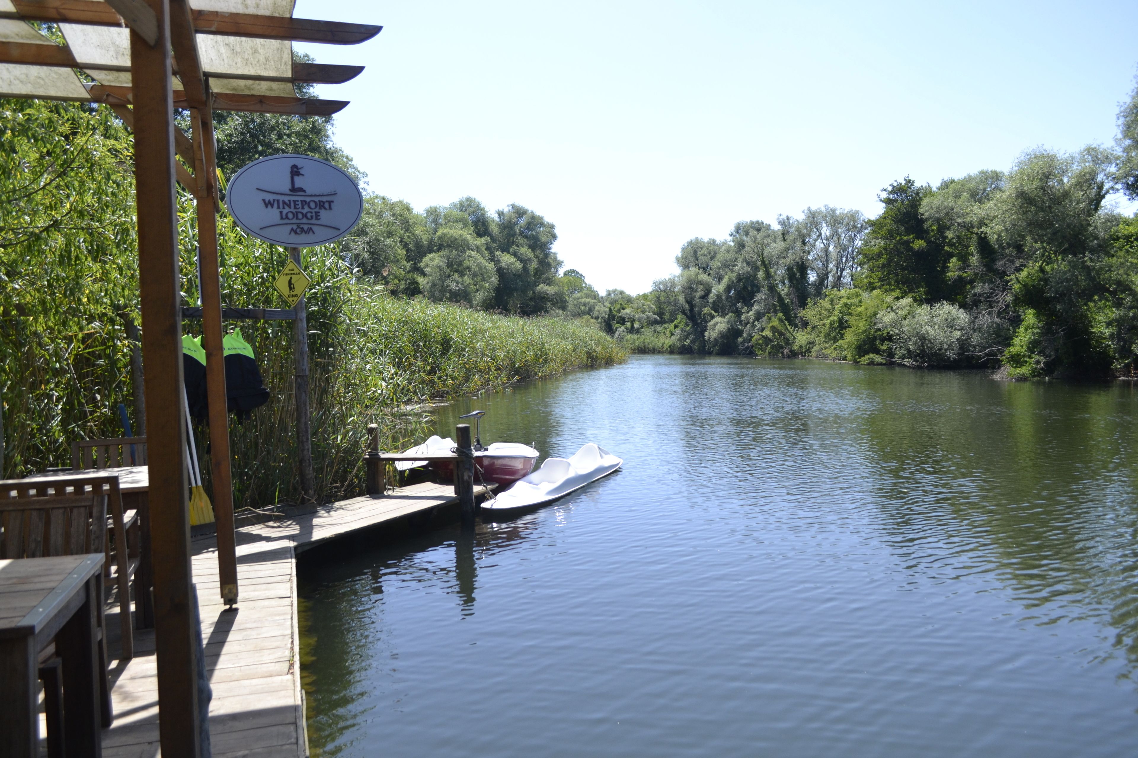 A wooden dock with a sign for Wineport Lodge, a boat docked at the pier, and a river surrounded by greenery.