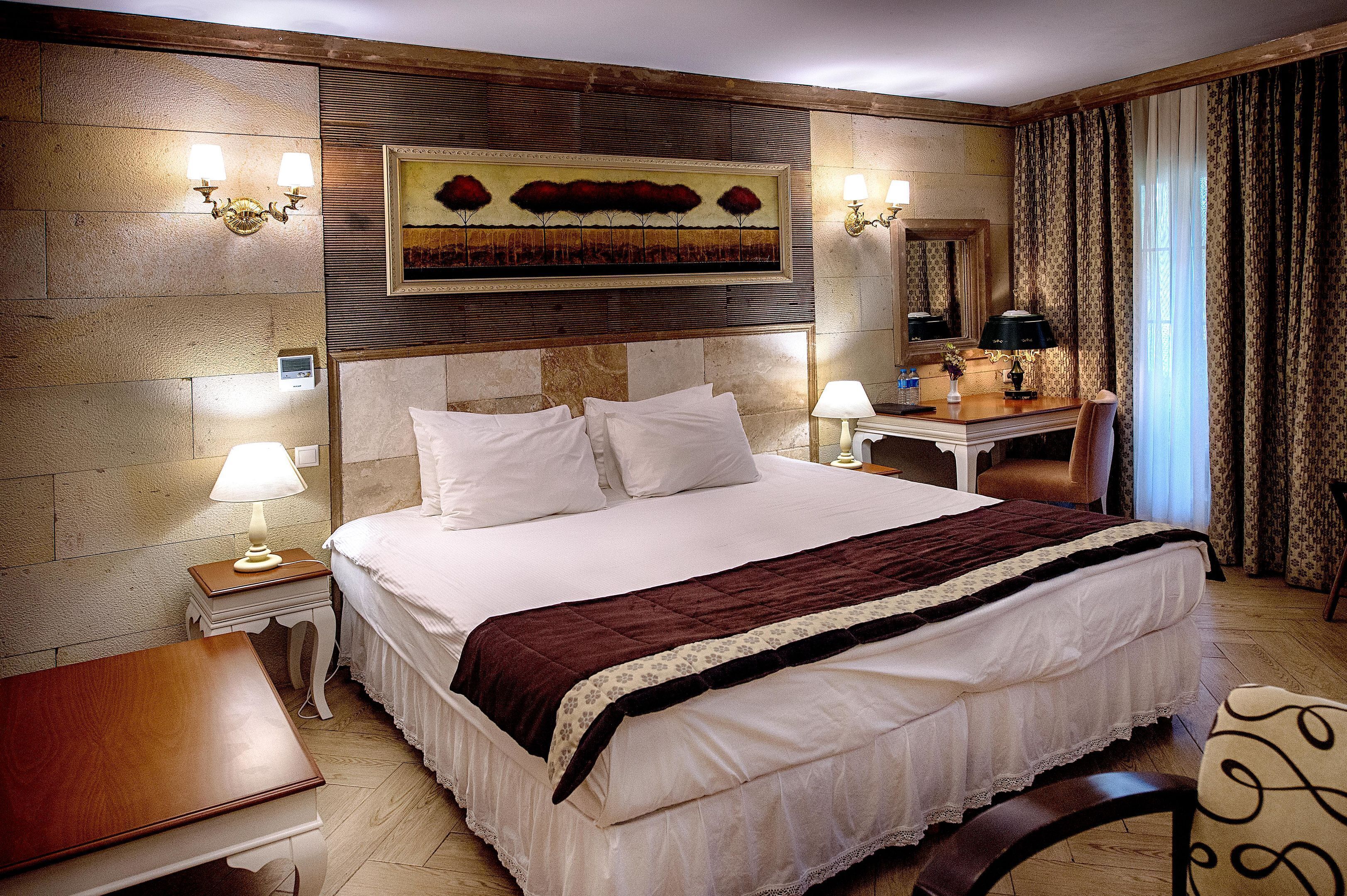 Habitación de hotel con una cama grande, dos mesitas de noche con lámparas, un escritorio con silla, un espejo y un cuadro en la pared.