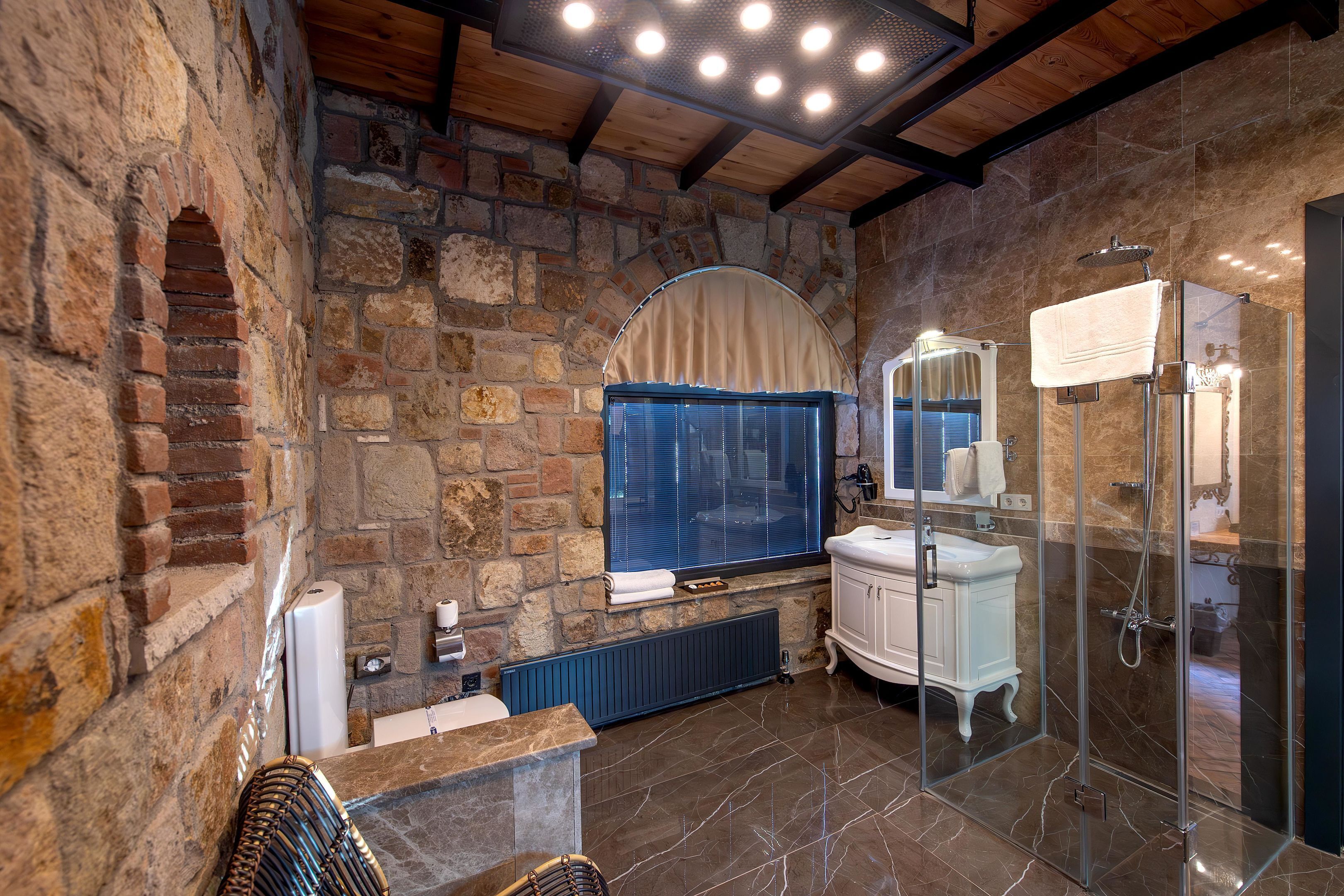 A bathroom with stone walls, a large window, a white sink, and a glass shower enclosure.