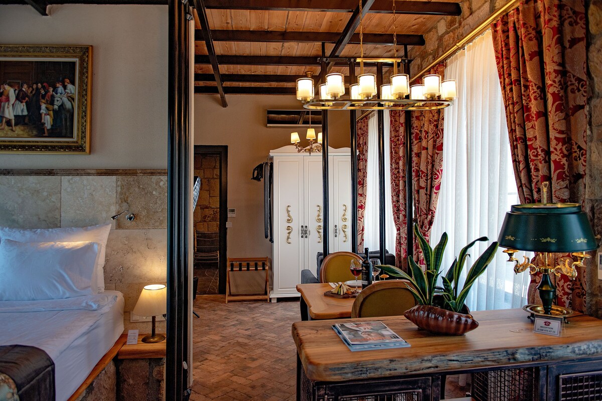 A hotel room with a bed, a table with a lamp, a chair, a potted plant, and a painting on the wall.