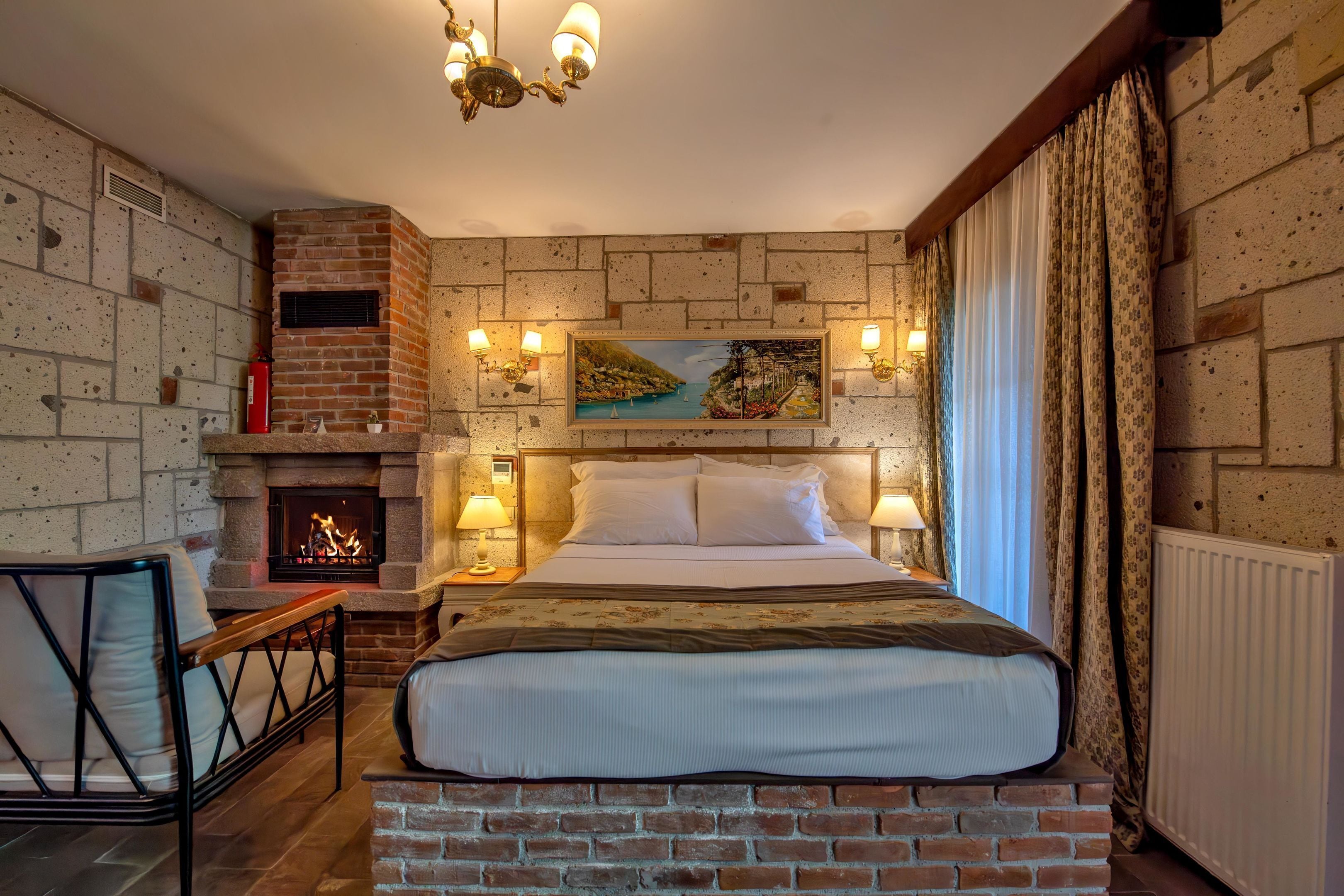 A bedroom with a brick fireplace, a bed with white linens, a nightstand with a lamp, and a painting on the wall.