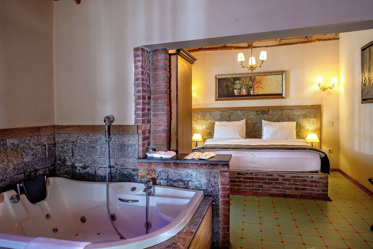 Quarto com uma banheira grande, cama com duas almofadas, mesa pequena com abajur e um quadro na parede.