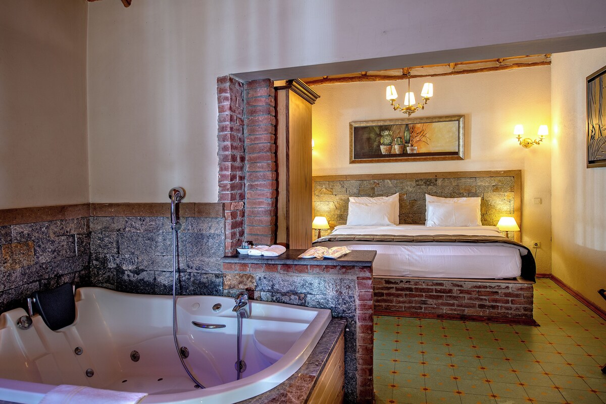 A room with a large bathtub, a bed with two pillows, a small table with a lamp, and a painting on the wall.