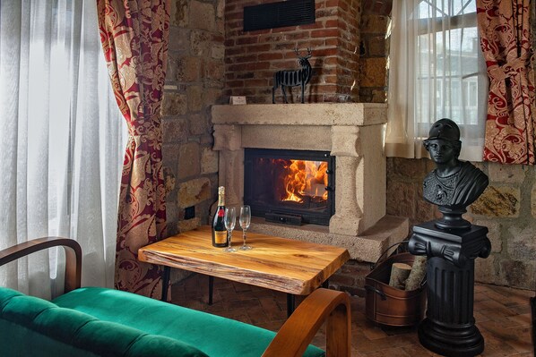 Una habitación acogedora con chimenea, una mesa con una botella y copas, un sofá verde y un busto.