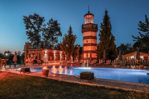 Zona de piscina con sillones de descanso y una fuente, rodeada de árboles y un edificio de piedra con torre.